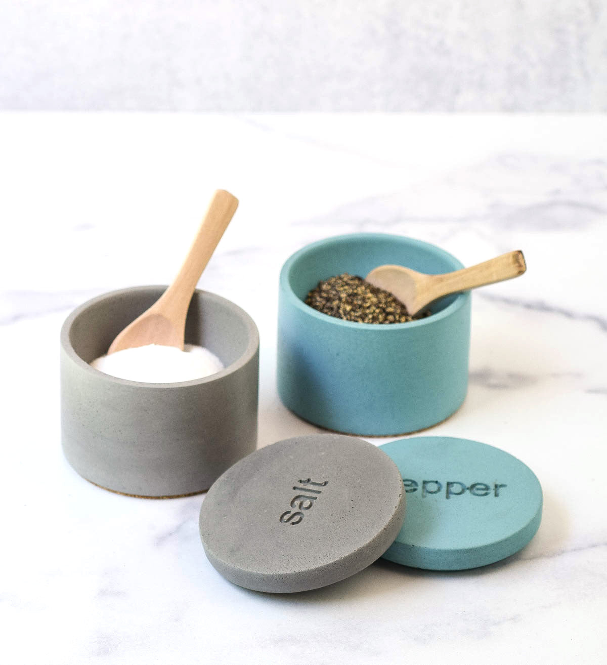 Gray salt cellar, lid off with spoon. Teal pepper cellar, lid off with spoon.