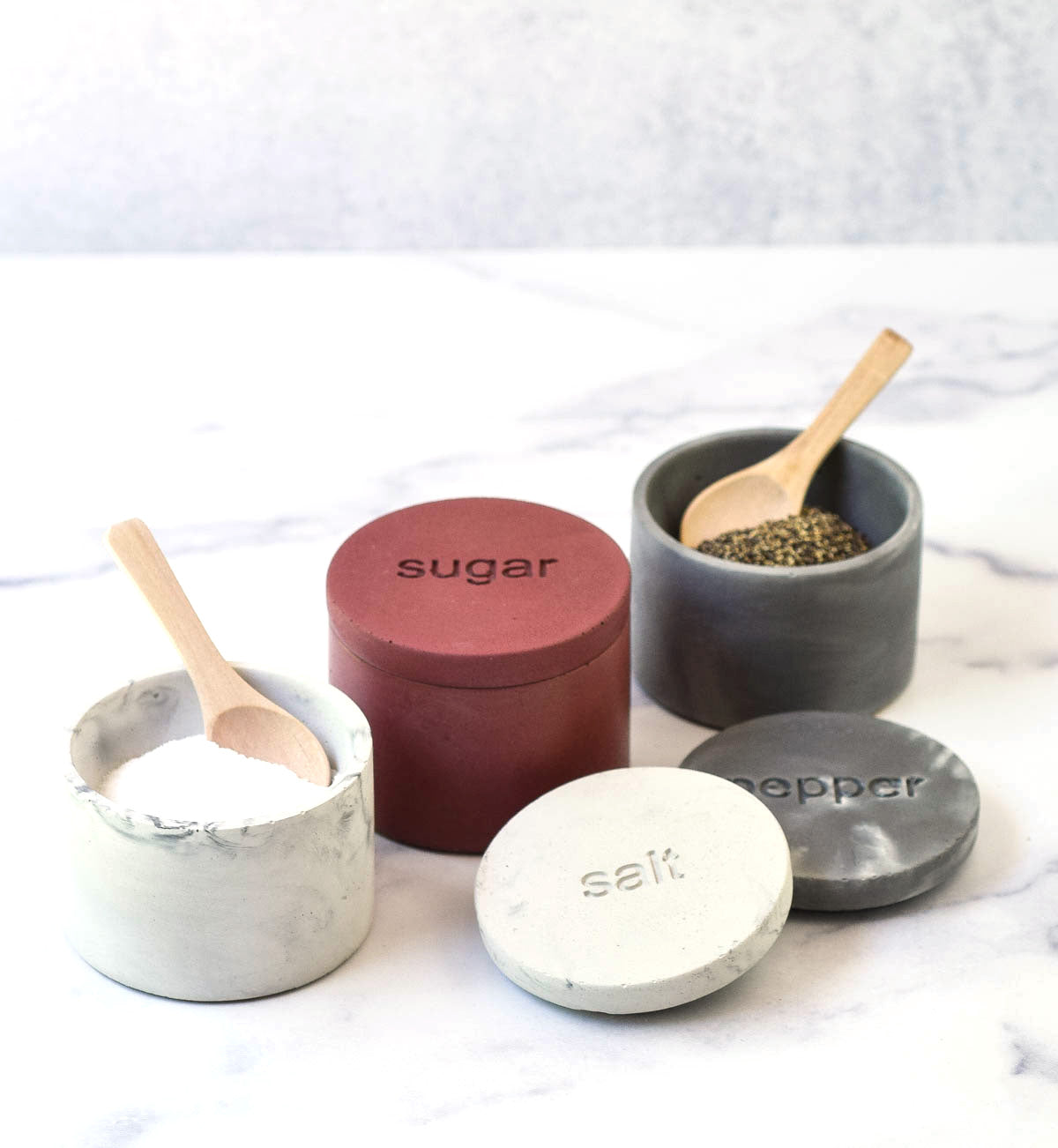 Marble salt cellar with lid off and spoon, plum sugar cellar with lid on, dark marble pepper cellar lid off with spoon.